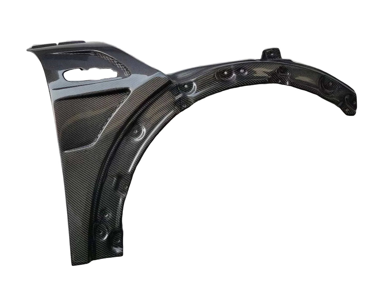 A carbon fiber automotive body panel or fender section with a glossy woven carbon pattern, photographed on a light gray ribbed surface.