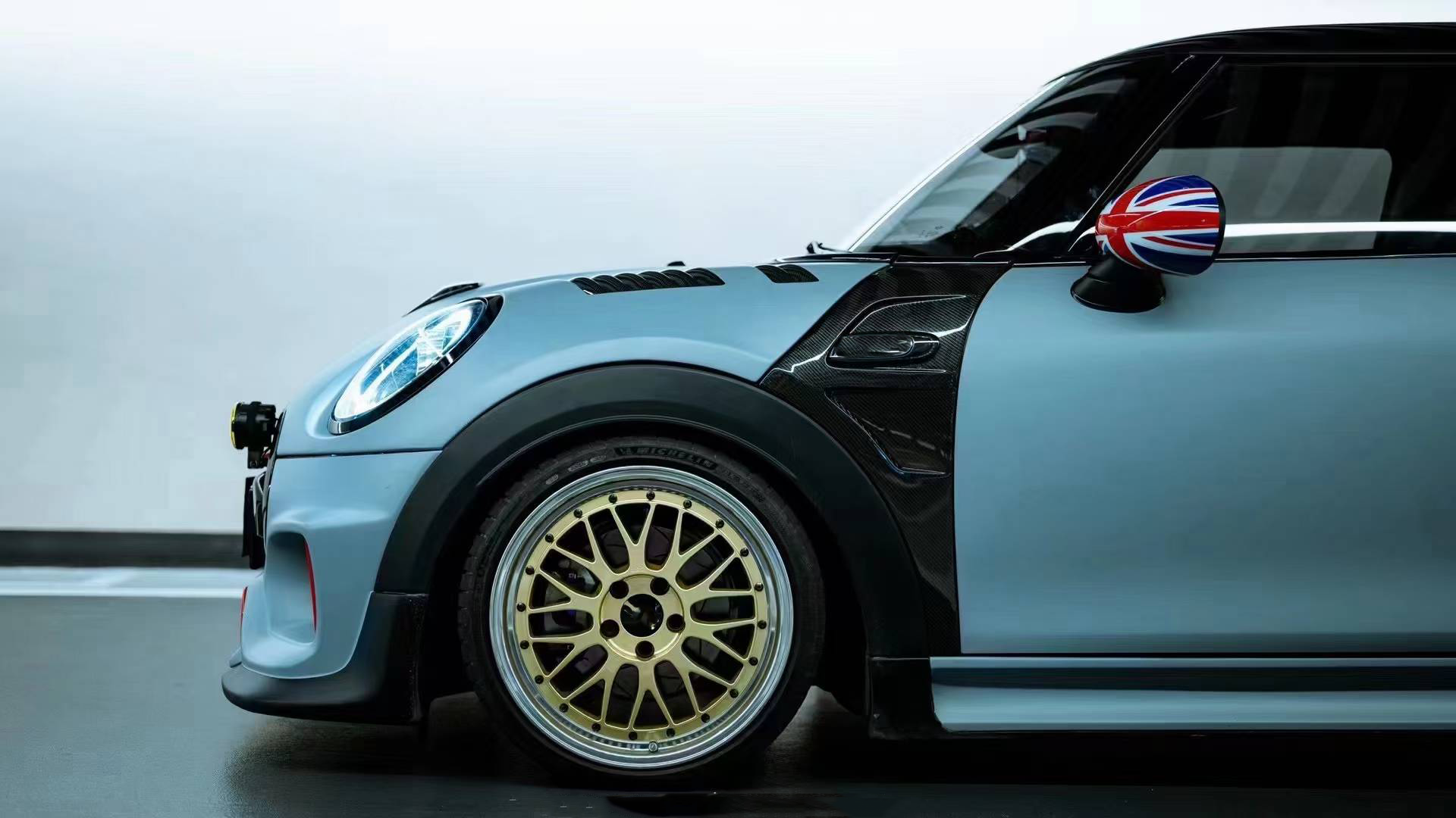 A light blue Mini Cooper with a widebody kit, black fender and wheel arches, gold multi-spoke wheels, and a Union Jack mirror, shown in a side profile against a gray background.
